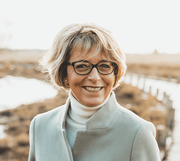 Elke teilt ihre Erfahung mit Emcomy. Blonde Frau Ü40 steht in der Natur und lächelt in die Kamera